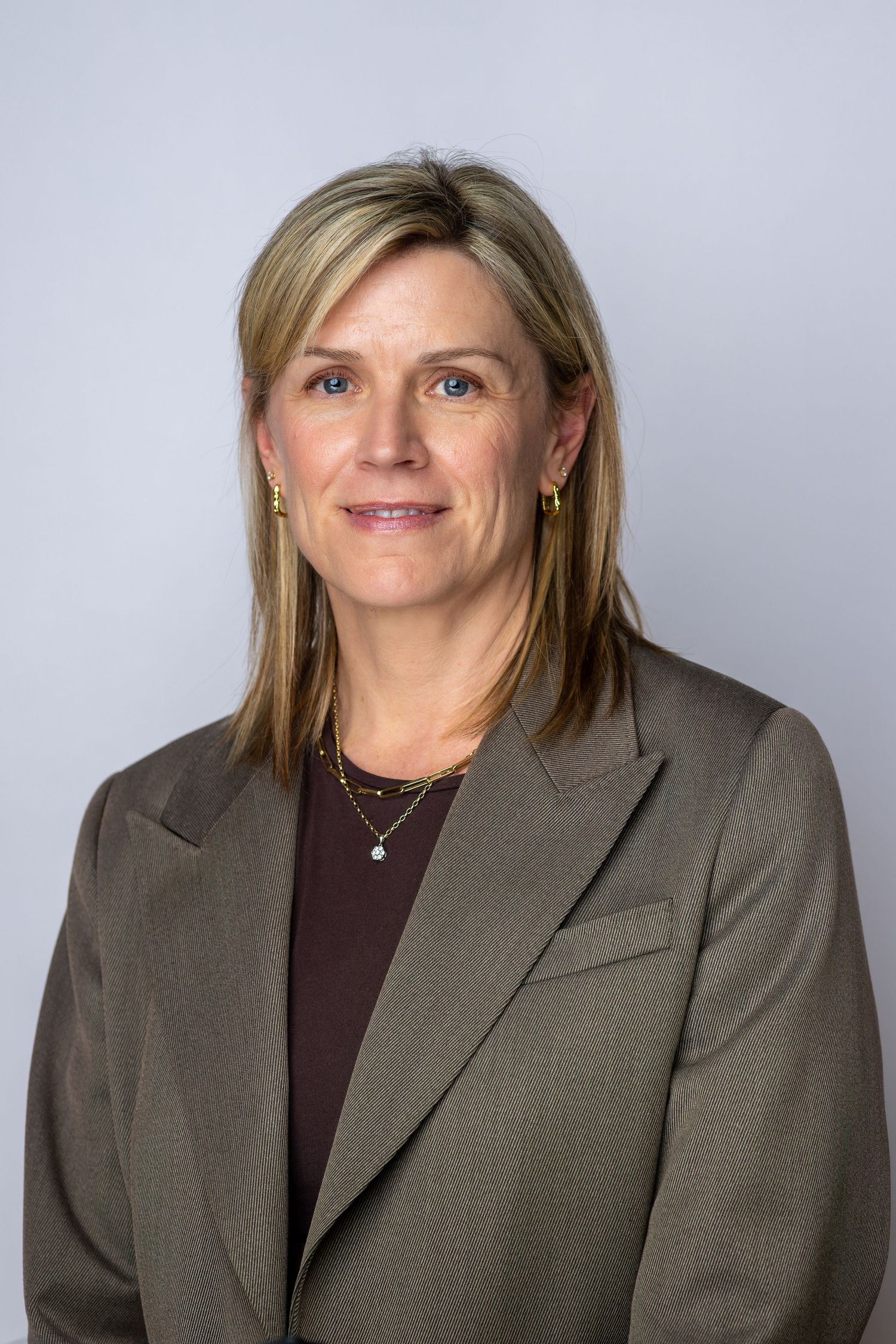 Professional headshot of a blonde woman in a olive blazer and burgundy top, wearing gold jewelry, against gray background.