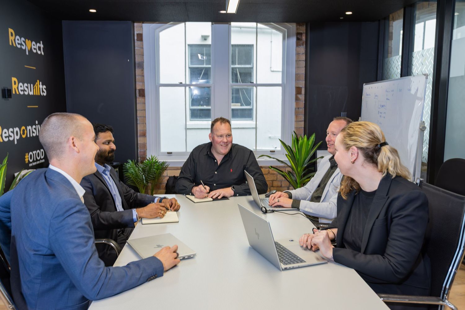 Five professionals in a modern meeting room having a business discussion around a white conference table with laptops.