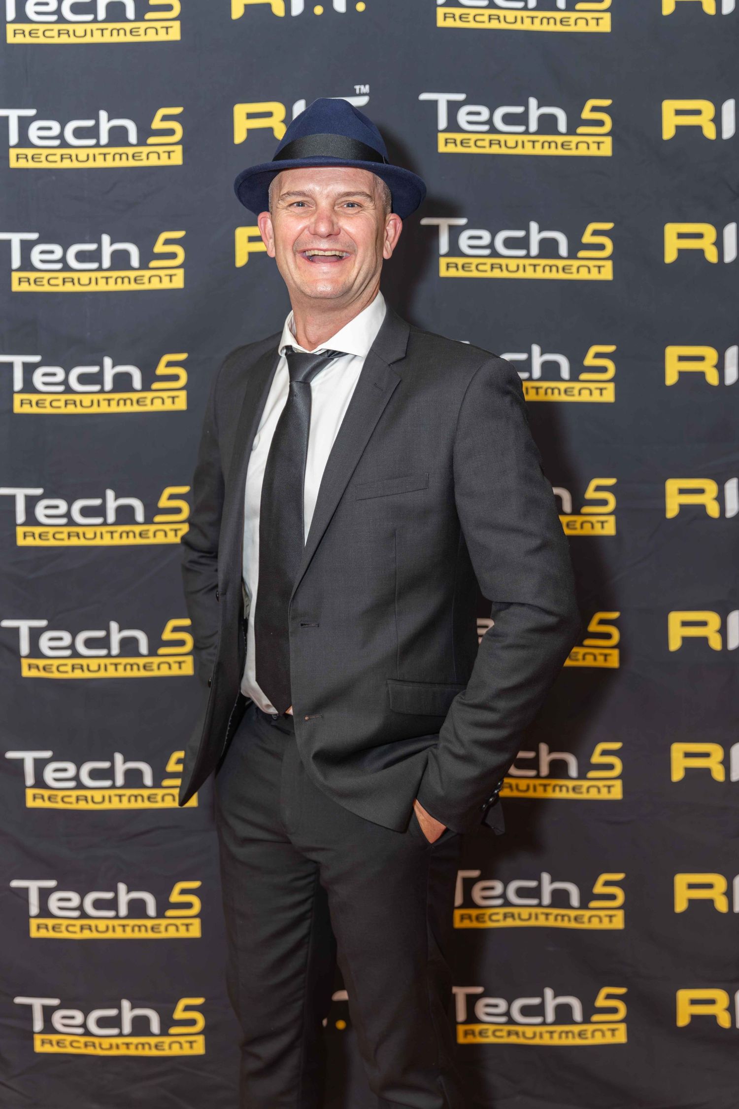 Man in dark suit and blue hat smiling at Tech5 Recruitment event backdrop.