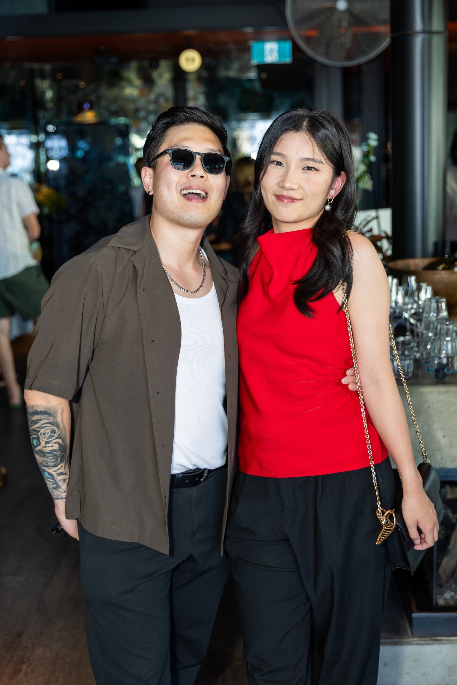 A man in sunglasses and a woman in red top pose together smiling at an indoor social event.