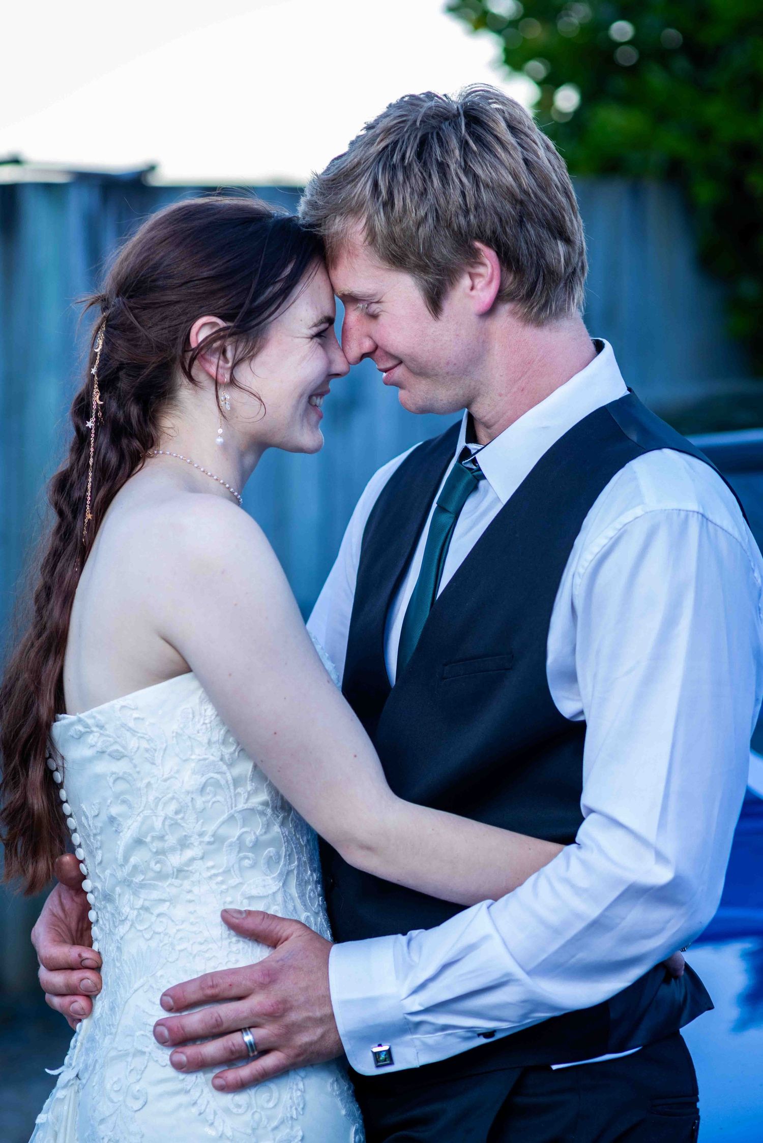 Bride and groom sharing a romantic forehead touch, smiling tenderly in wedding attire outdoors at dusk.