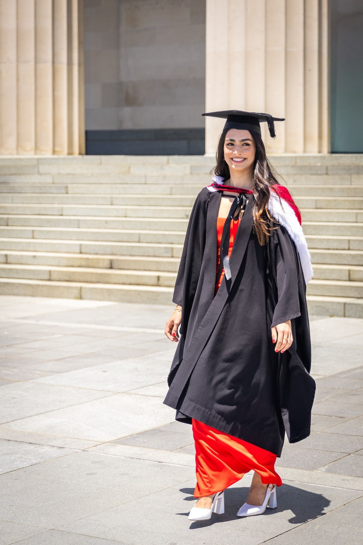 A smiling female graduate in black cap and gown with red sash stands on steps of a university building.