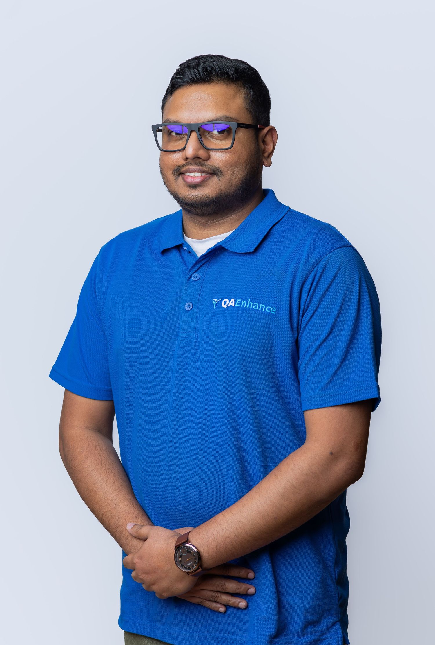 Man in blue QAEnhance polo shirt posing professionally against light gray background, wearing glasses and a watch.