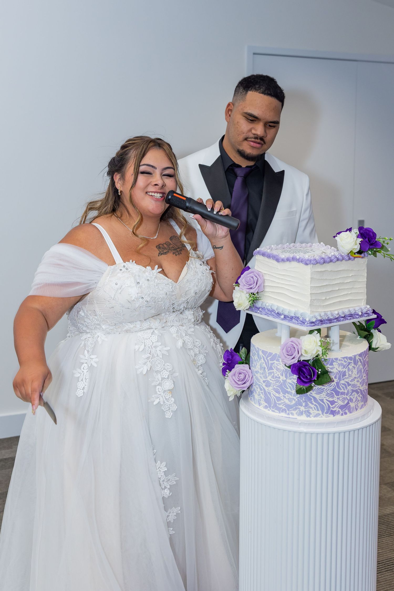 Bride and groom cutting a three-tier purple and white wedding cake, bride holding microphone in bridal gown.