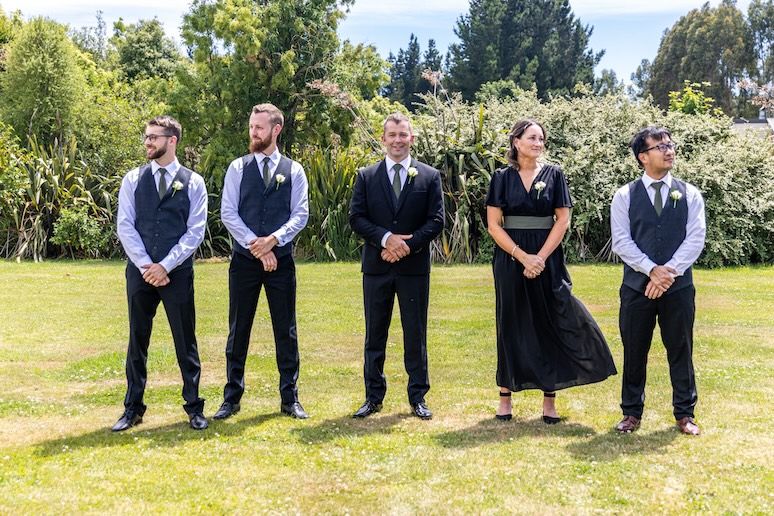 Wedding party of five standing on green lawn, groomsmen in black suits with vests and a woman in elegant black dress.