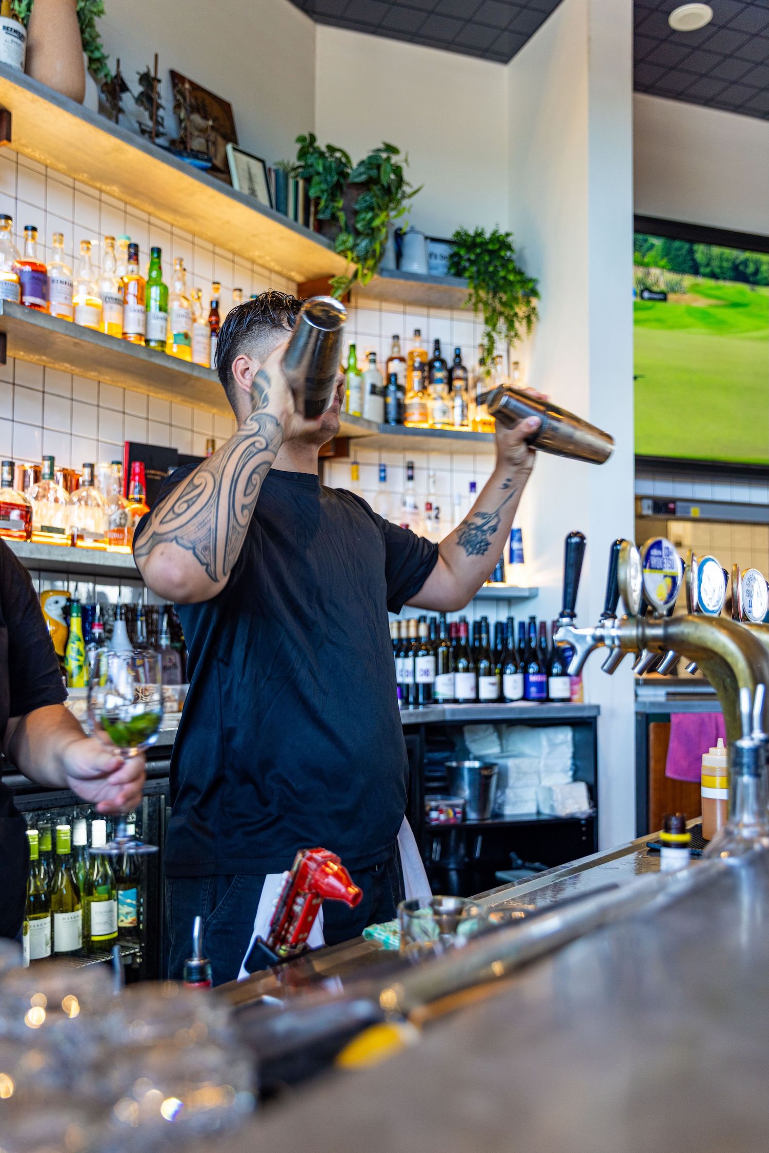 A bartender in black attire mixes drinks at a well-stocked bar with illuminated shelves and bottles on display.