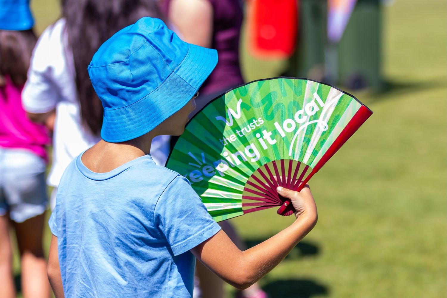 Attendees at an outdoor event hold green folding fans while sitting under blue umbrellas on a sunny day.