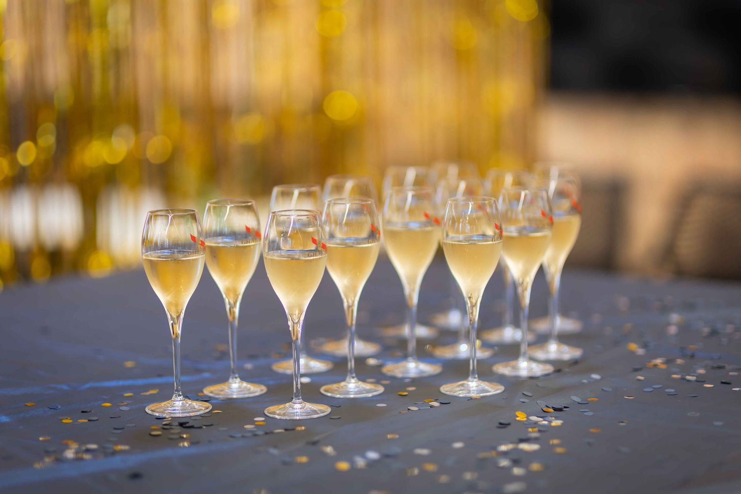 Rows of elegant champagne flutes filled with golden sparkling wine on a table with gold tinsel backdrop.