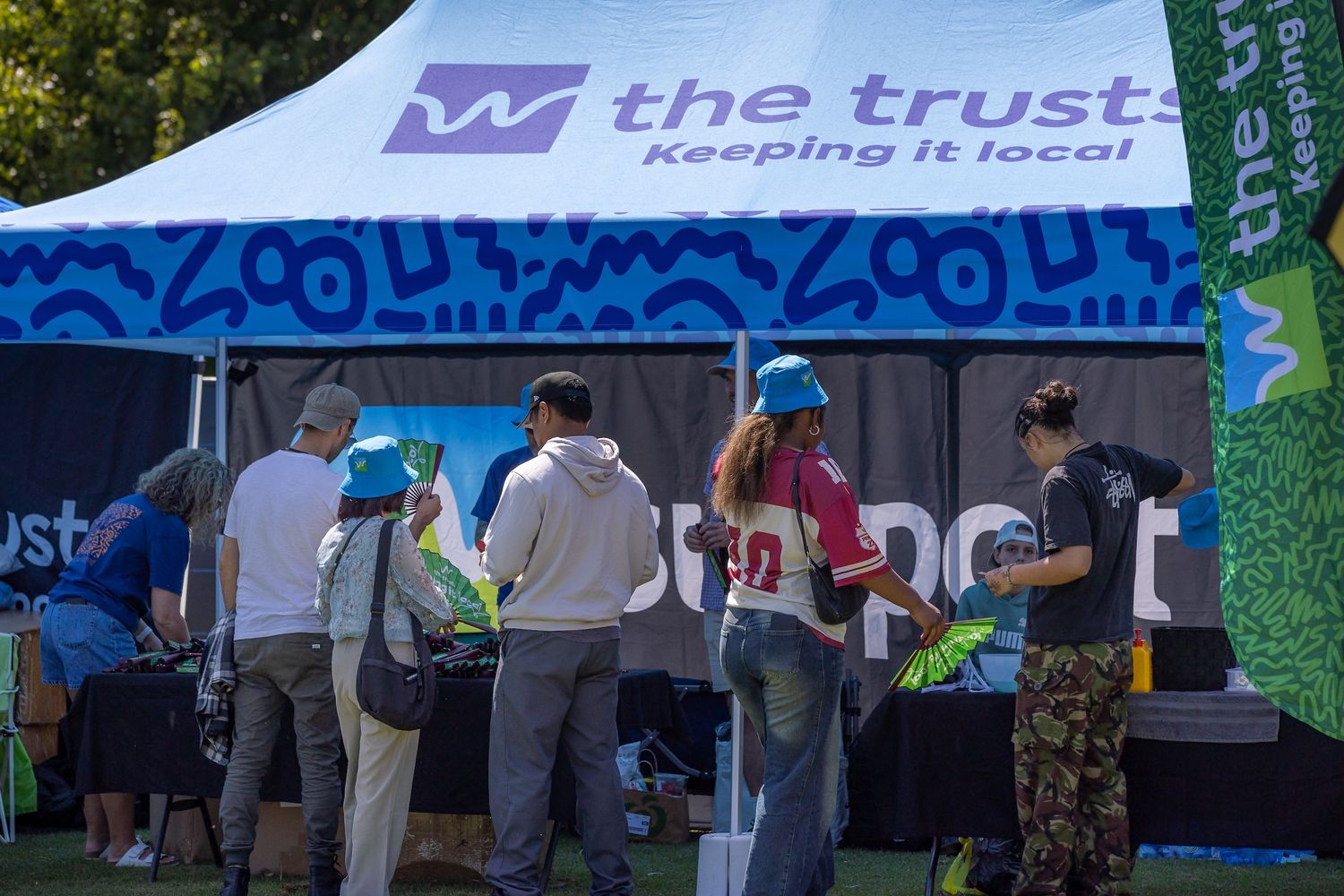 Outdoor event setup with white tents and banners displaying 'The Trusts' branding on a sunny day.