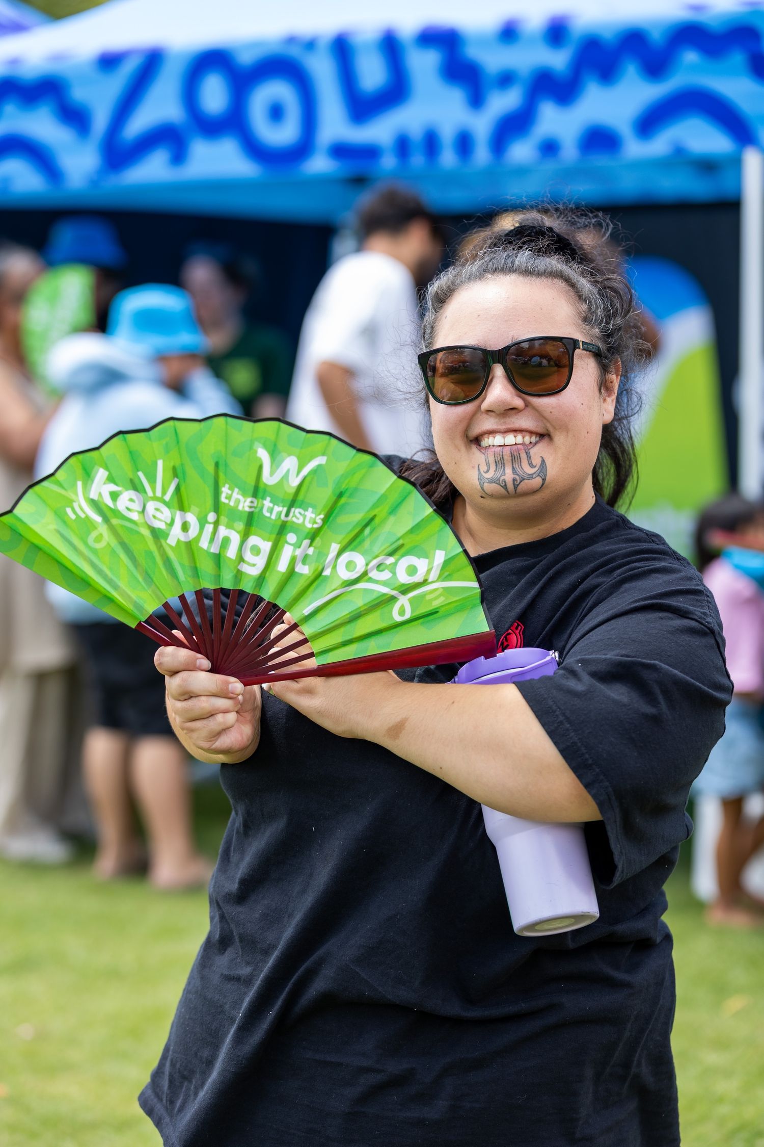 People holding green promotional fans at outdoor event with blue tent in background.