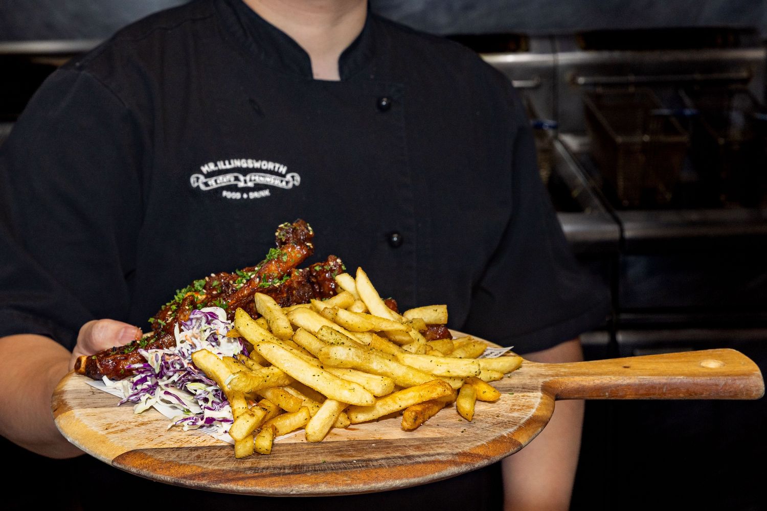 A wooden serving board with crispy french fries and coleslaw being presented by someone in a black chef coat.