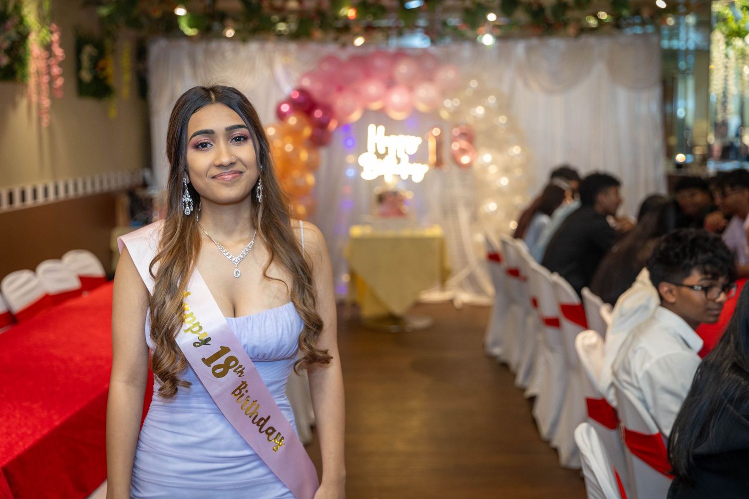 Young woman in white dress with 18th birthday sash smiles at elegant party venue with balloon arch.