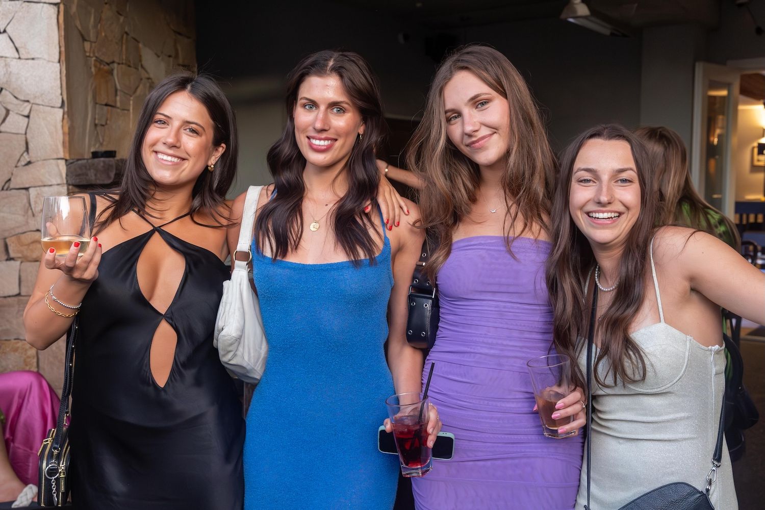 Four women in elegant dresses smile together at a social gathering, holding drinks in a stylish venue.
