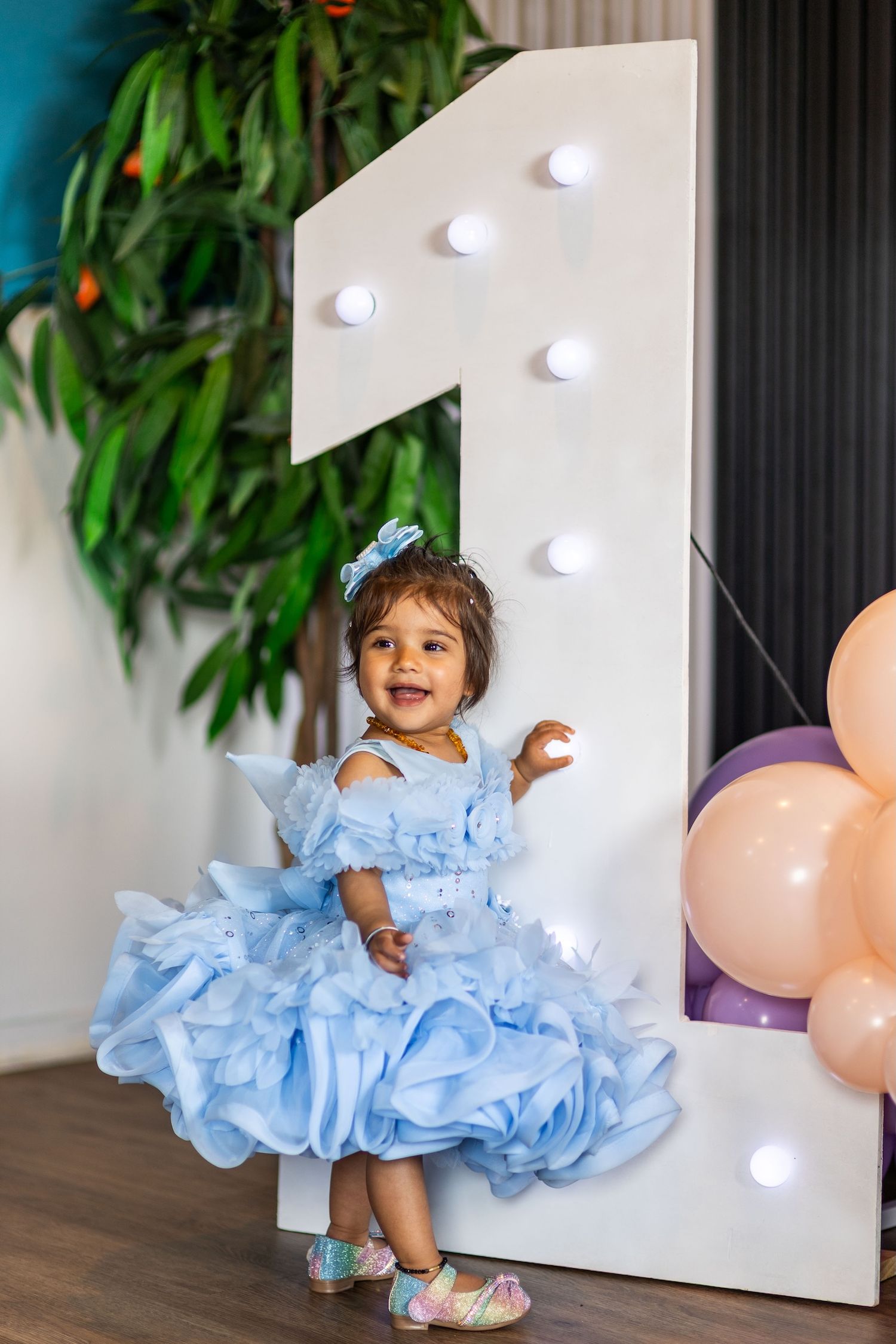 Smiling toddler girl in blue ruffle dress celebrating 1st birthday with balloons and illuminated number one backdrop.