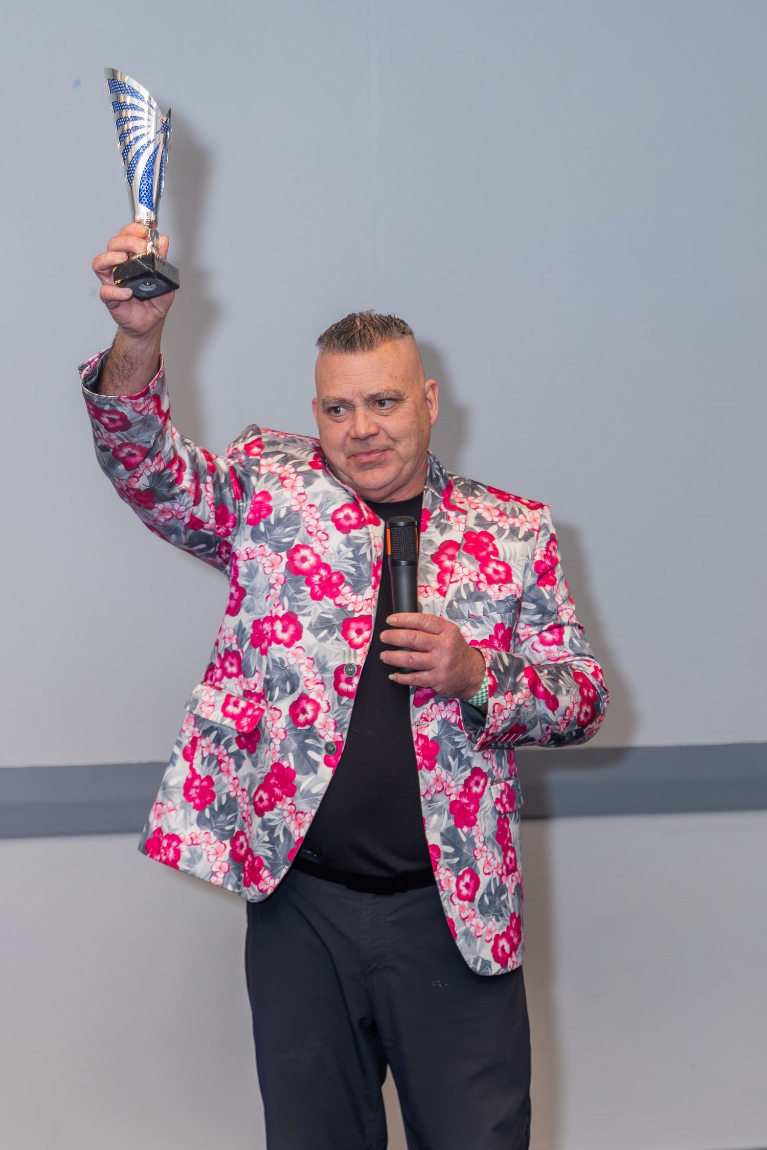 Man in floral blazer holding trophy award aloft while gripping microphone at awards ceremony.