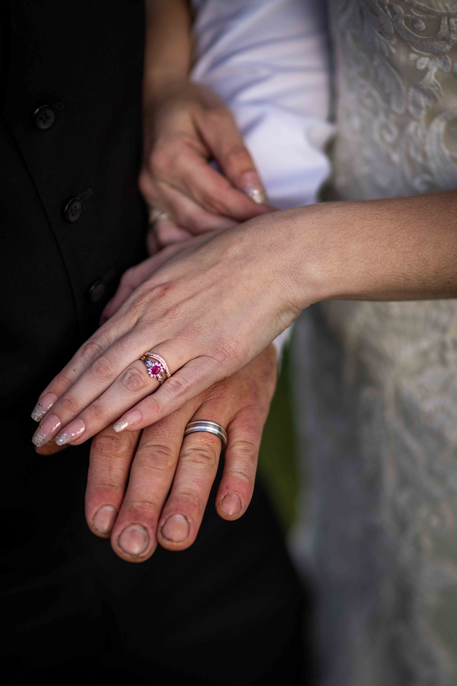 Newlywed couple displaying wedding rings, bride wearing pink gemstone ring, groom with silver band, hands clasped together.