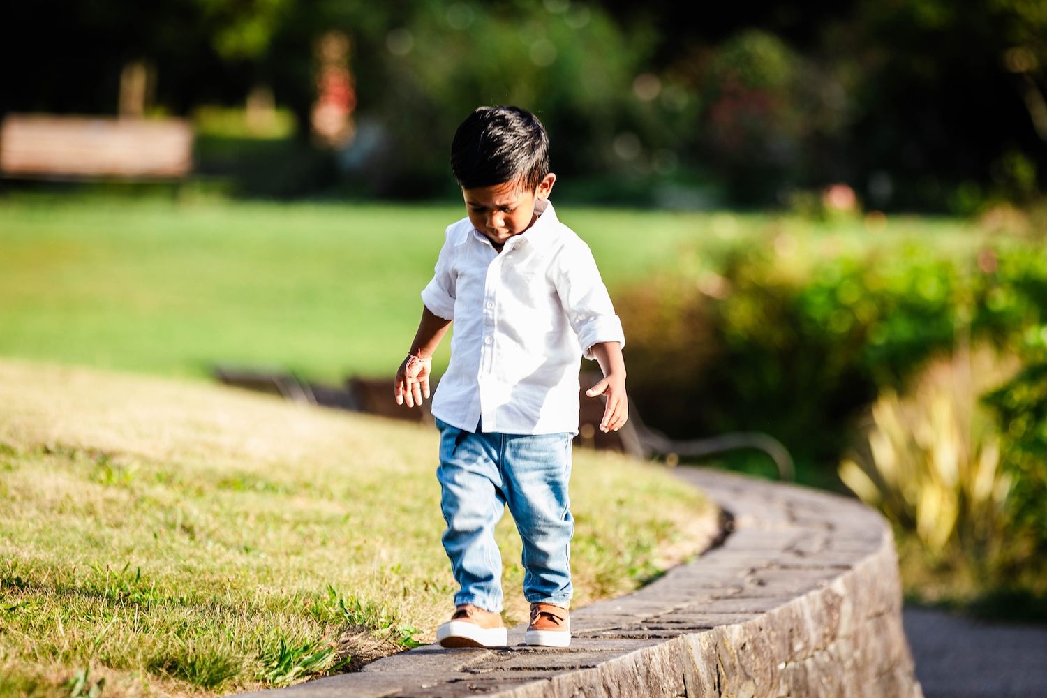 Young boy in white shirt and jeans balancing on a stone path in a sunny green park.