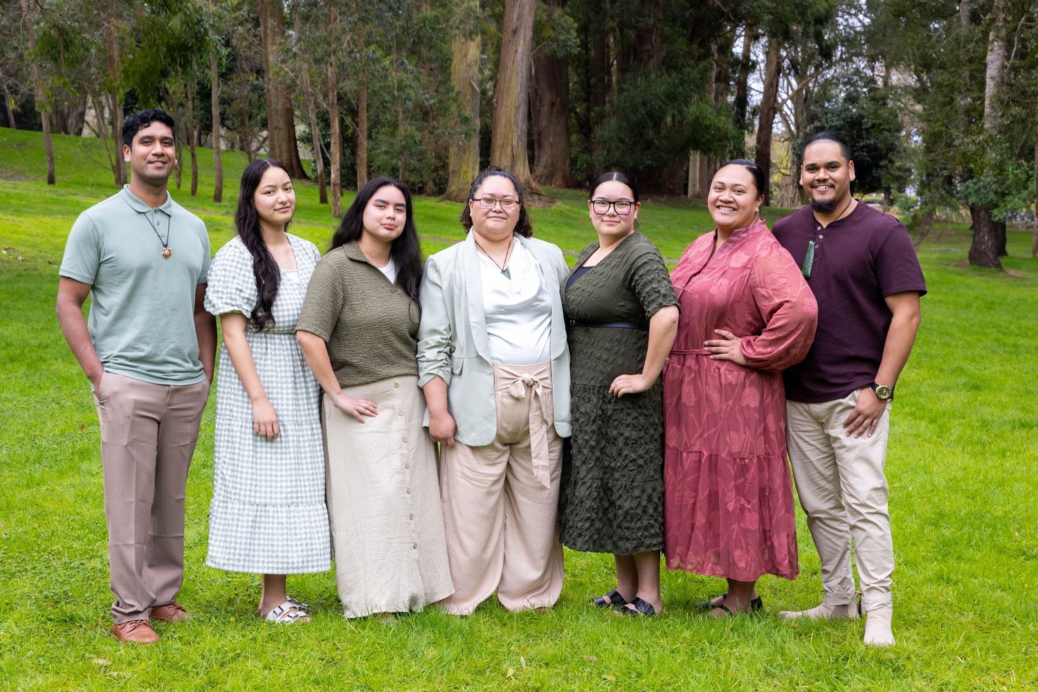Seven people smiling and posing together outdoors on green grass surrounded by tall trees.