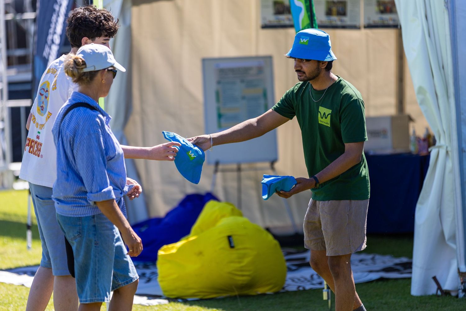 Group of people gathered at outdoor festival with tents and yellow beanbag visible in background.