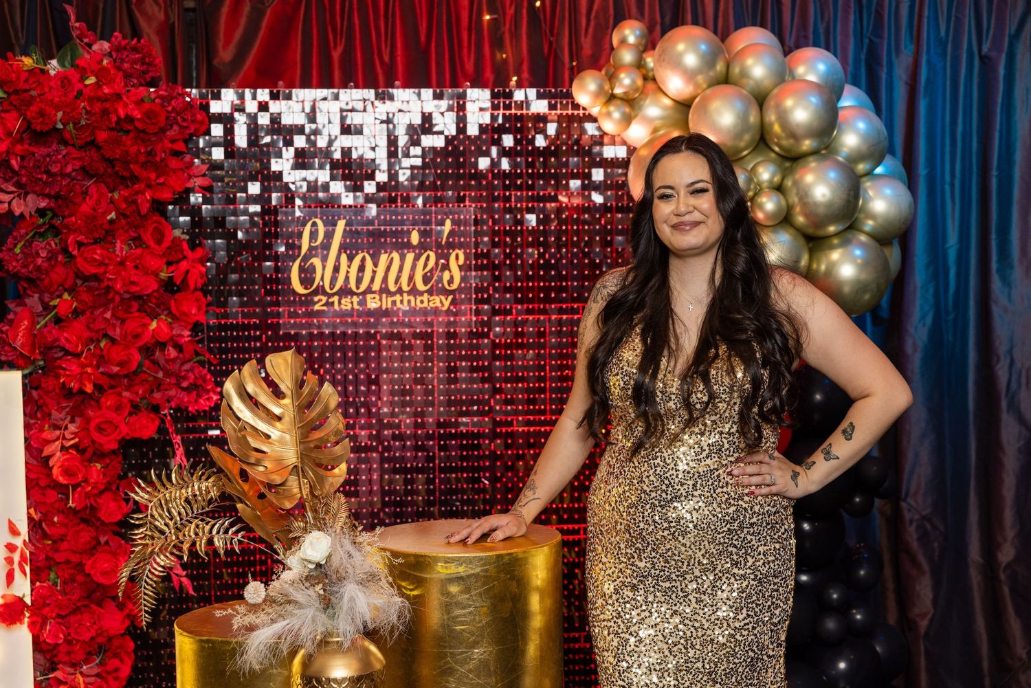 Woman in gold sequin dress poses at Ebonie's birthday party with balloon arch and red floral decorations.