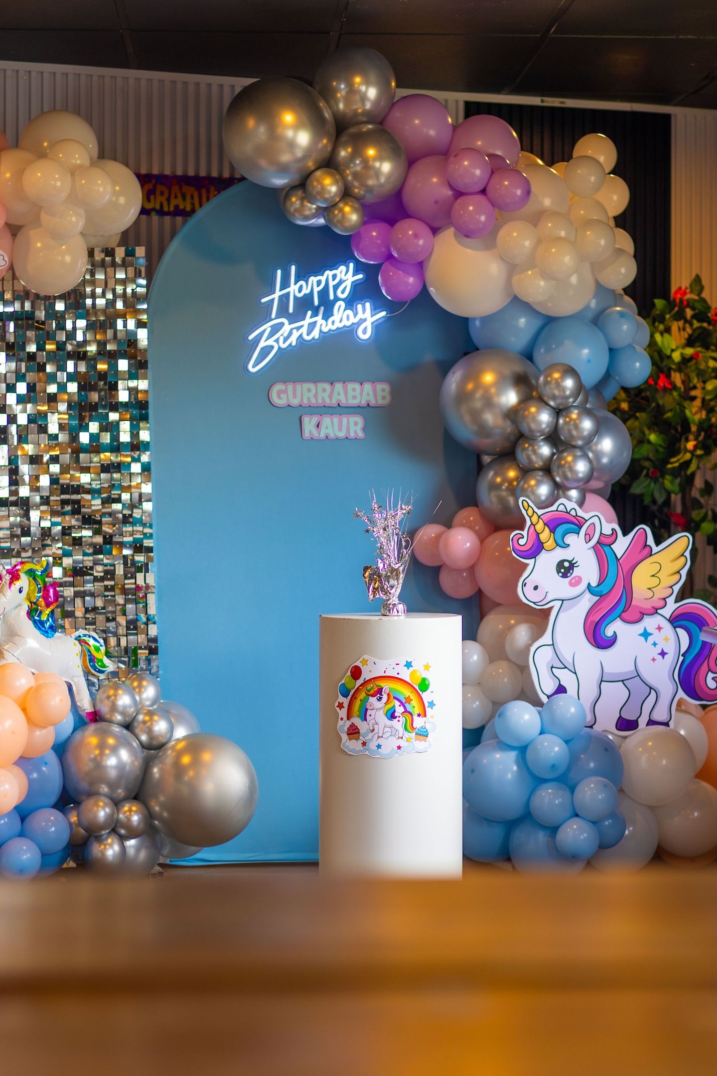 Unicorn-themed birthday party setup with balloon arch, neon Happy Birthday sign, and decorative pedestal.