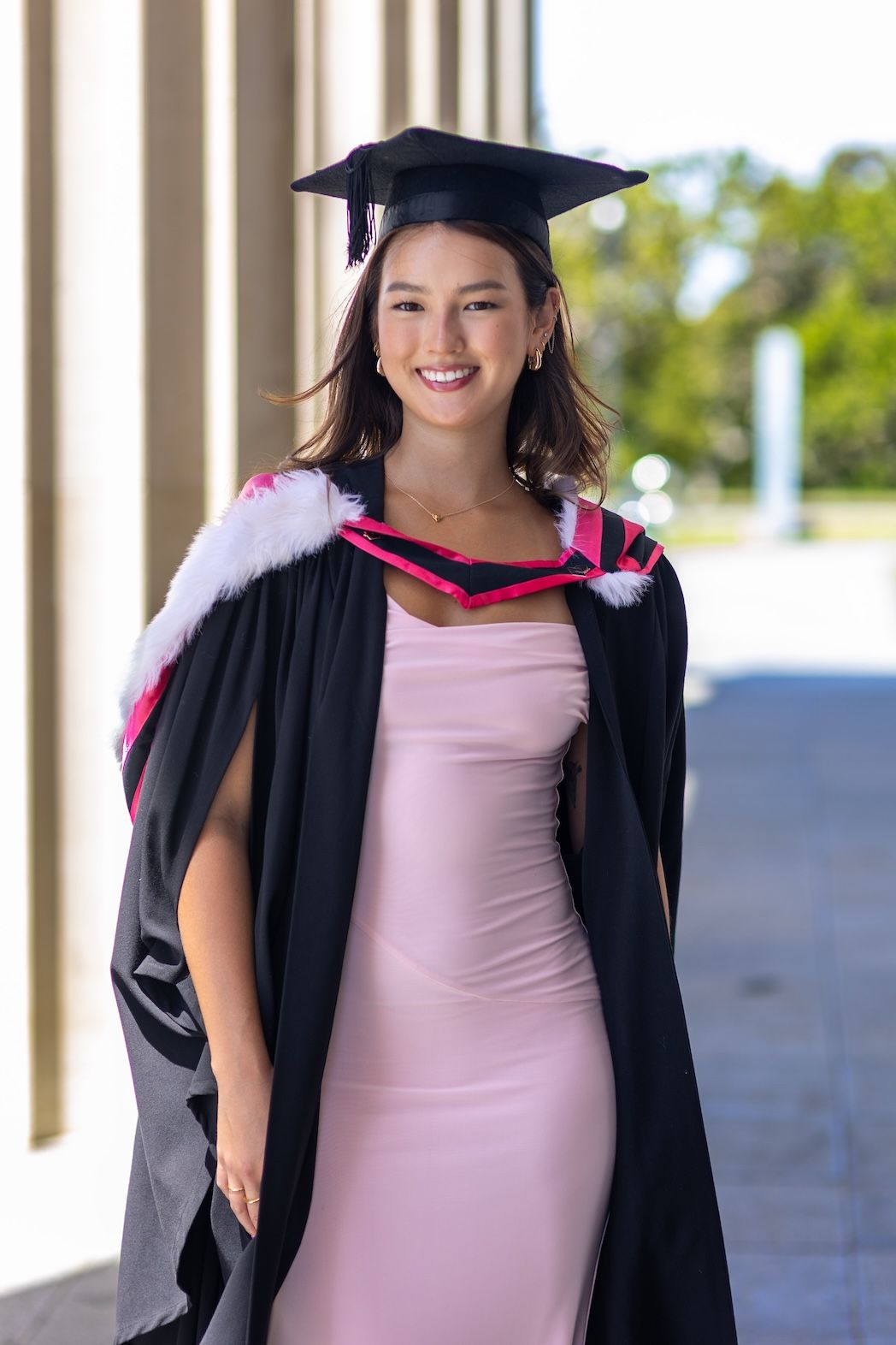 Smiling young woman in black graduation gown and cap with pink hood, wearing a pink dress outdoors.