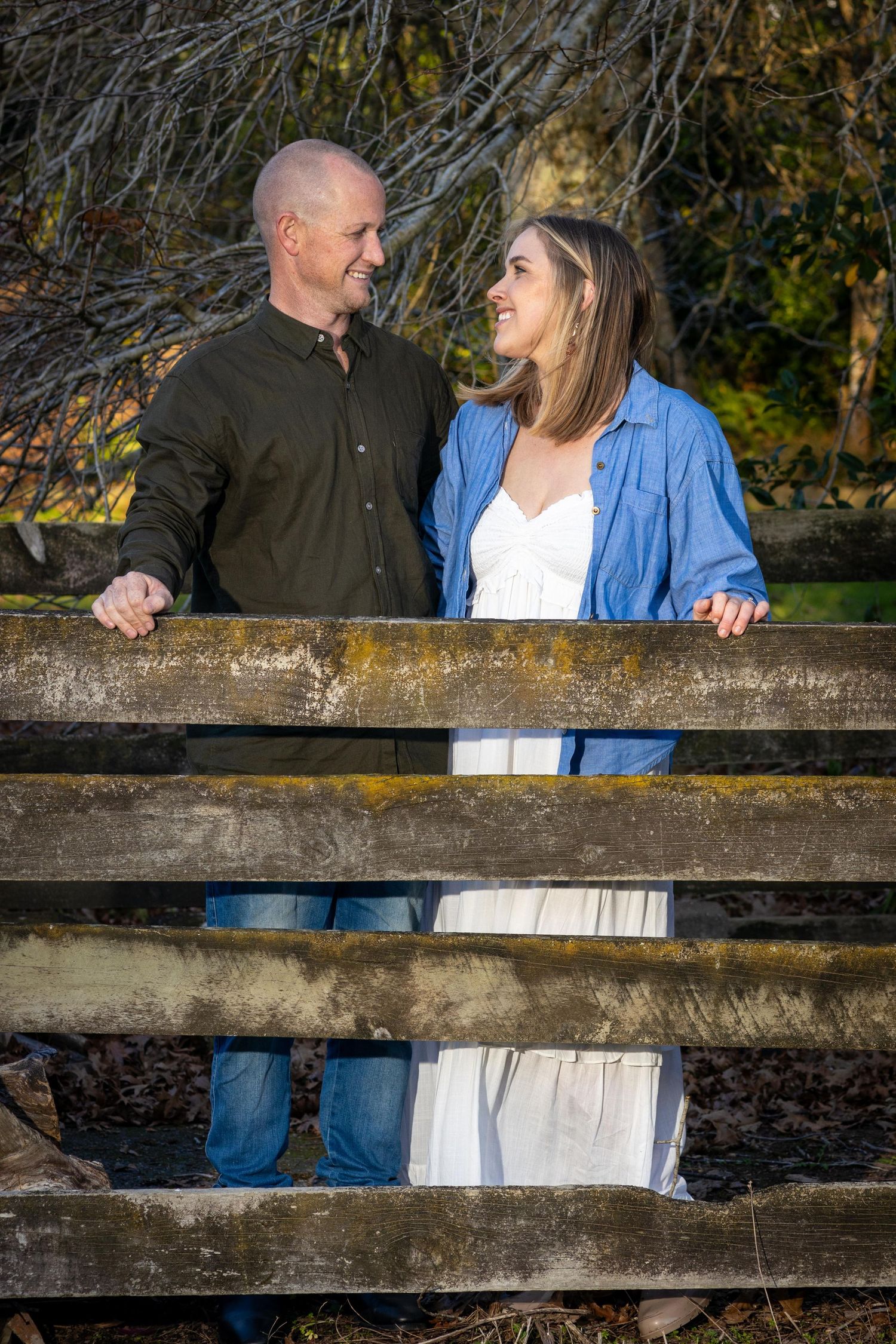 A couple smiles at each other while standing behind a rustic wooden fence in an autumn outdoor setting.