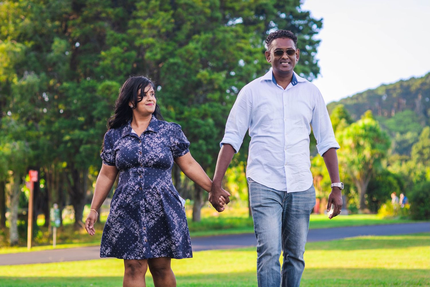 Couple holding hands walking in a lush green park on a sunny day, wearing casual summer outfits.