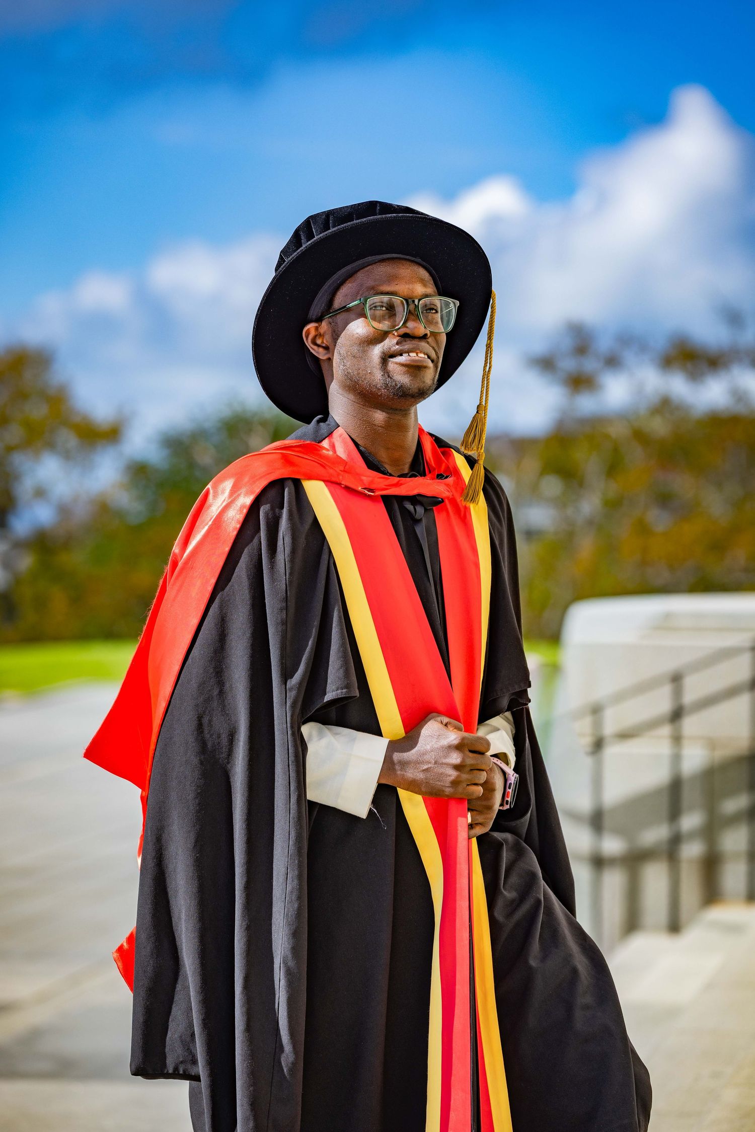 A proud Black man in doctoral graduation gown with red and gold hood and mortarboard hat smiles outdoors on a sunny day.