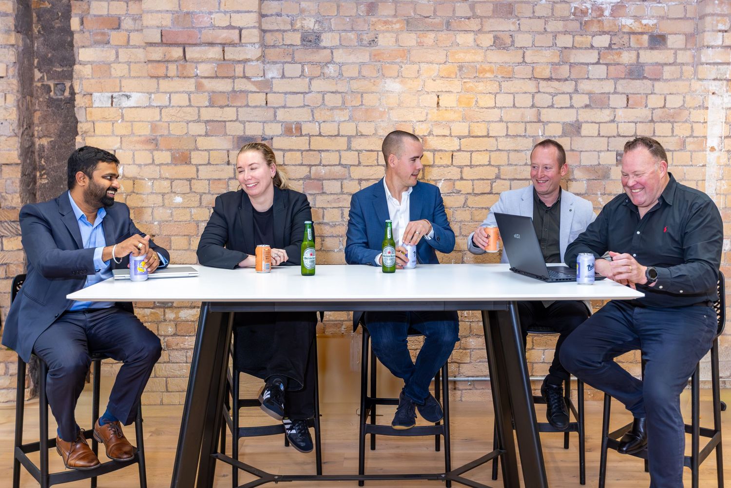 Five business professionals sitting at a white table in a modern brick-walled office, smiling and collaborating together.