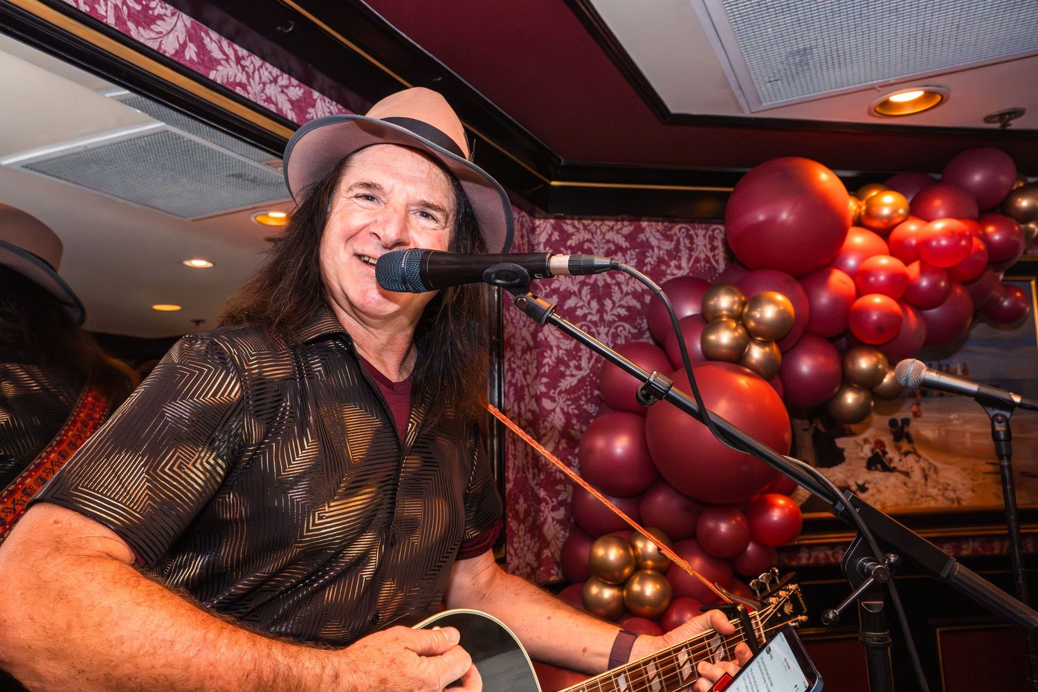 Smiling male guitarist performing live with microphone, wearing brown hat, colorful balloons in background.