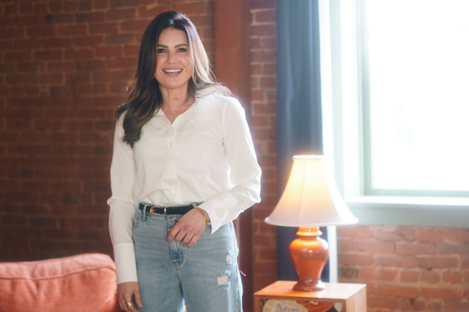 Woman in white shirt and jeans smiling in a brick-walled room with warm lamp light.