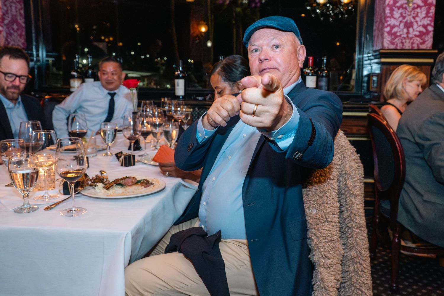 Man in blue jacket and cap points at camera while dining at an elegant restaurant with wine glasses.