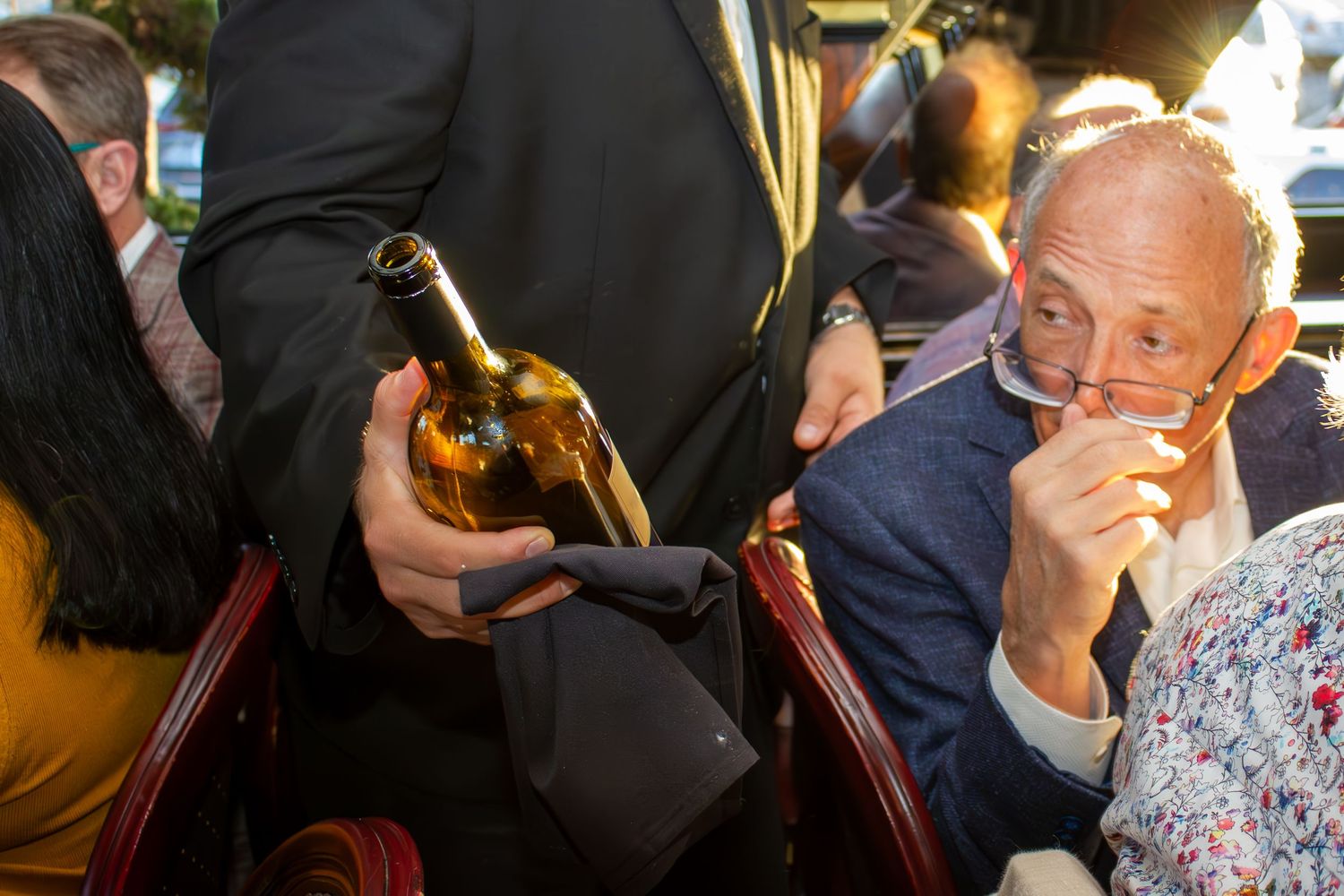 An elderly man examines a wine bottle offered by a waiter at an elegant dinner event.