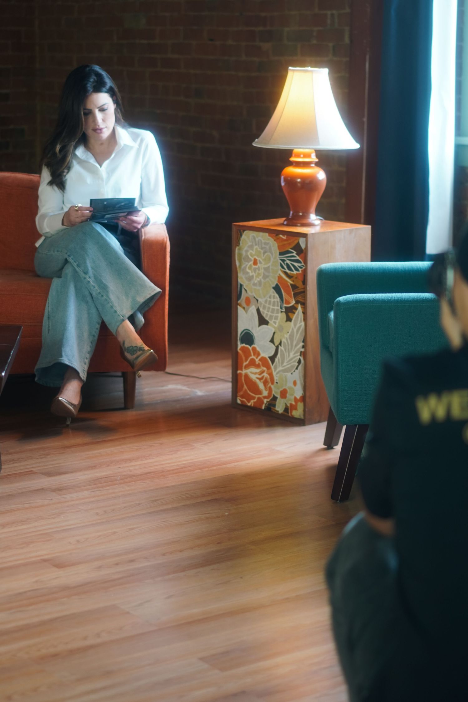 Woman in white blouse sits in orange chair using tablet beside floral side table with glowing lamp.