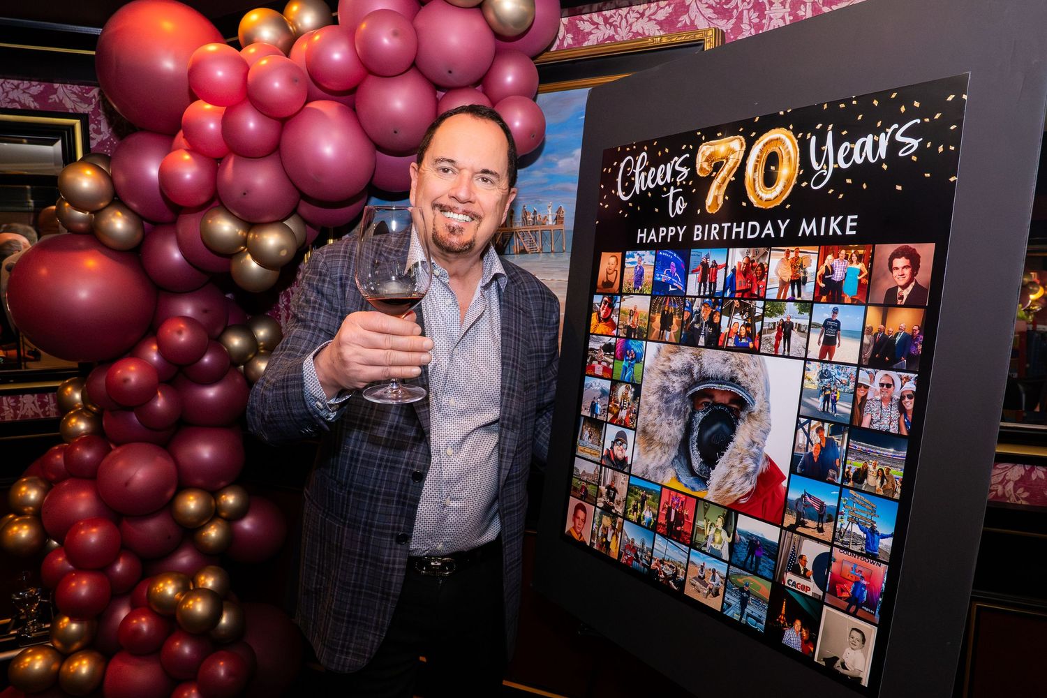 Man celebrating 70th birthday party holding wine glass beside balloon arch and photo collage display.