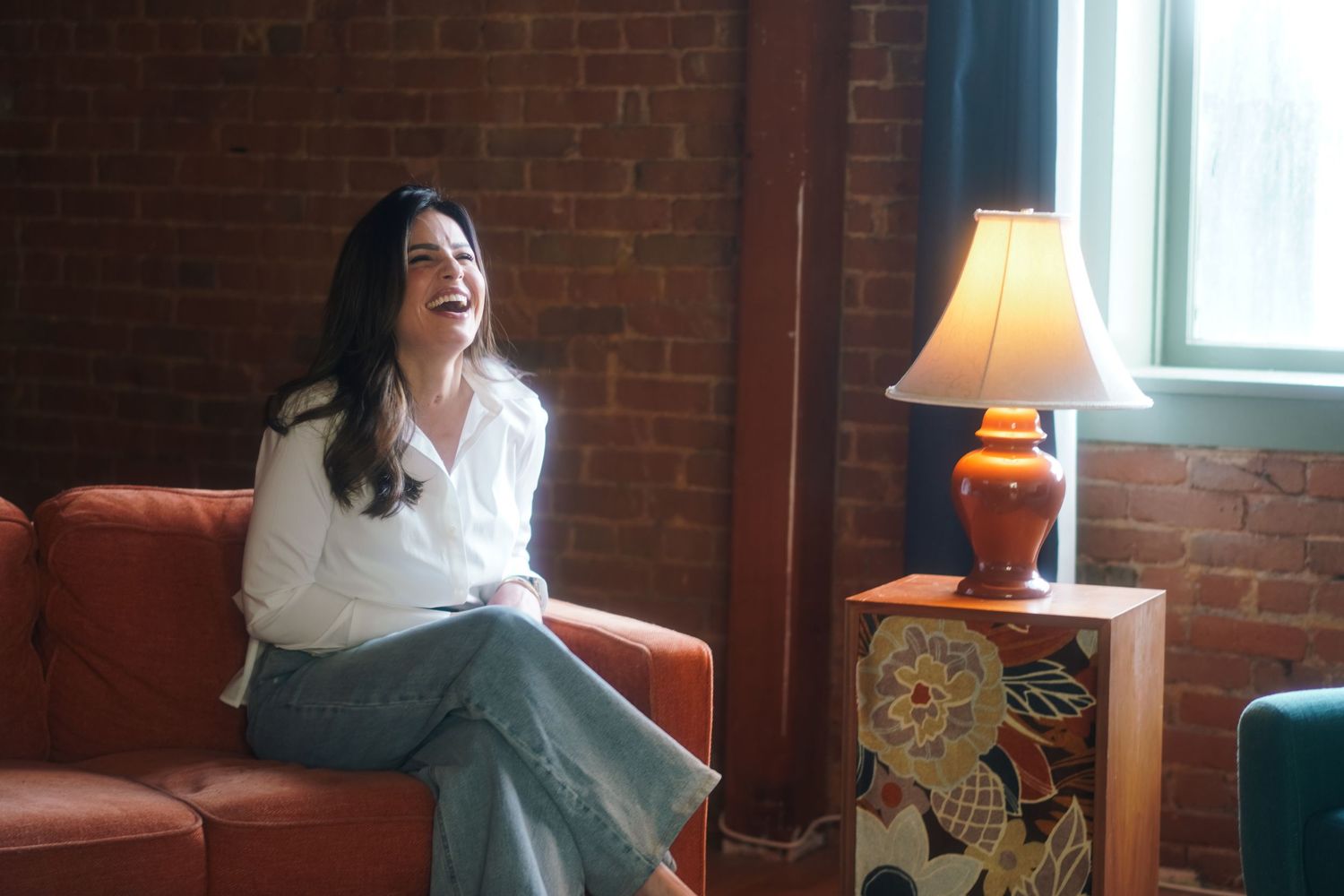 Woman in white blouse and jeans laughing on red sofa near brick wall and decorative lamp.