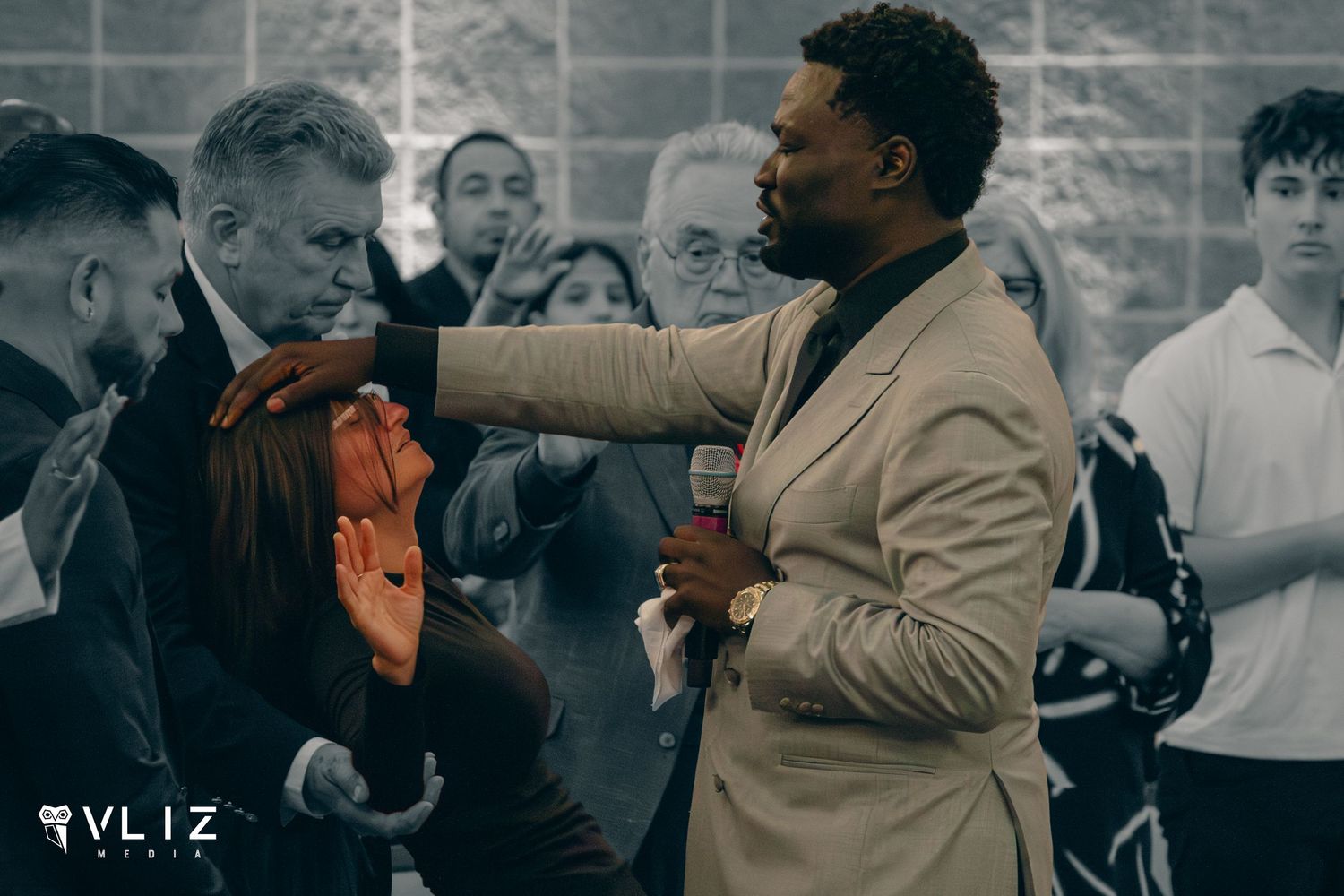 A man in a beige suit lays hands on a woman during a prayer service at a church gathering.