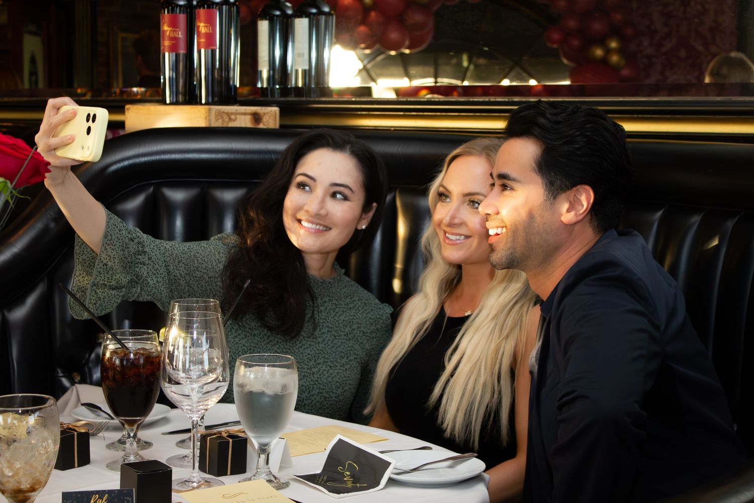 Three friends taking a selfie at an elegant restaurant dinner table with wine glasses and dim lighting.
