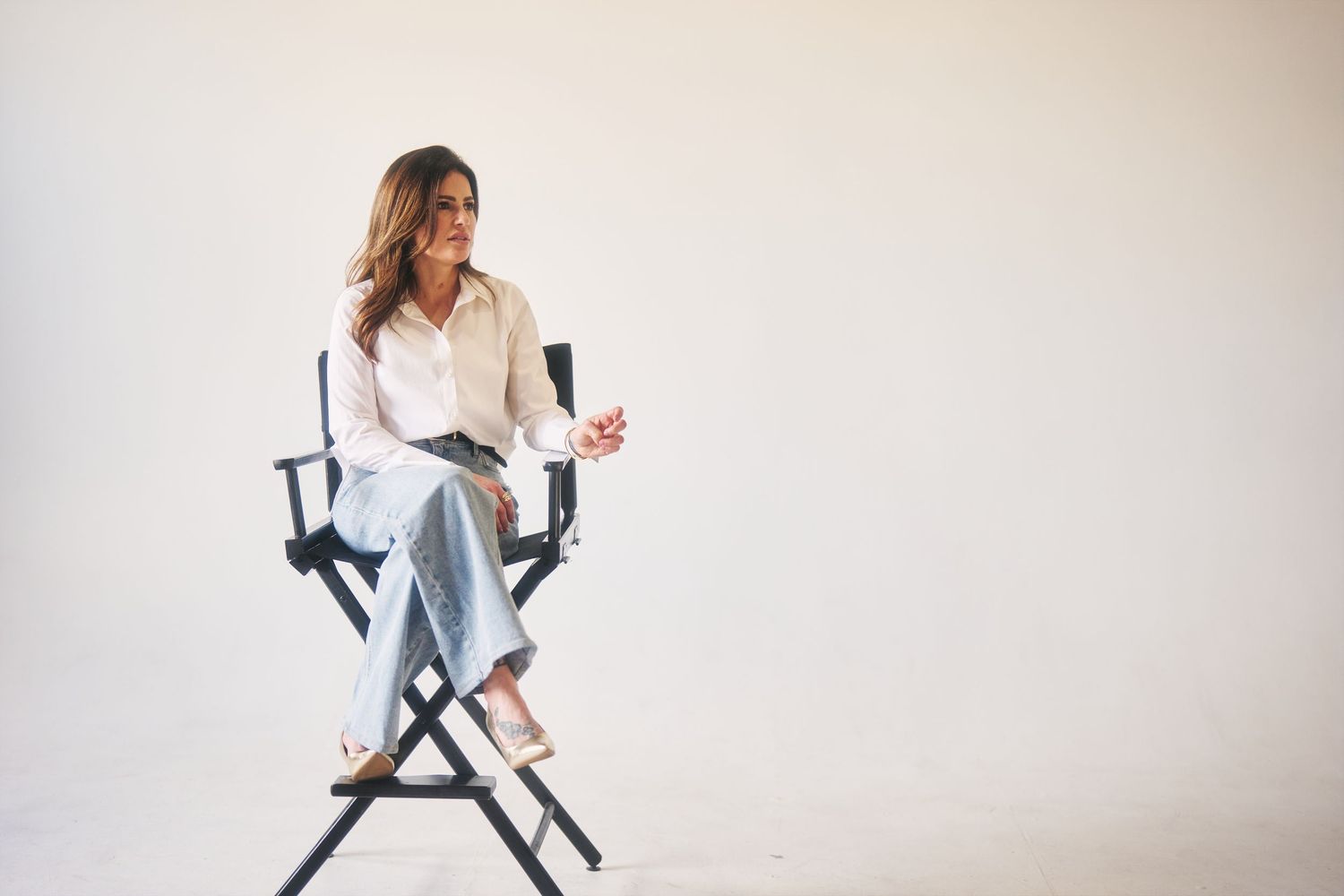 Woman in white shirt and jeans sitting relaxed in a director's chair against a white background.