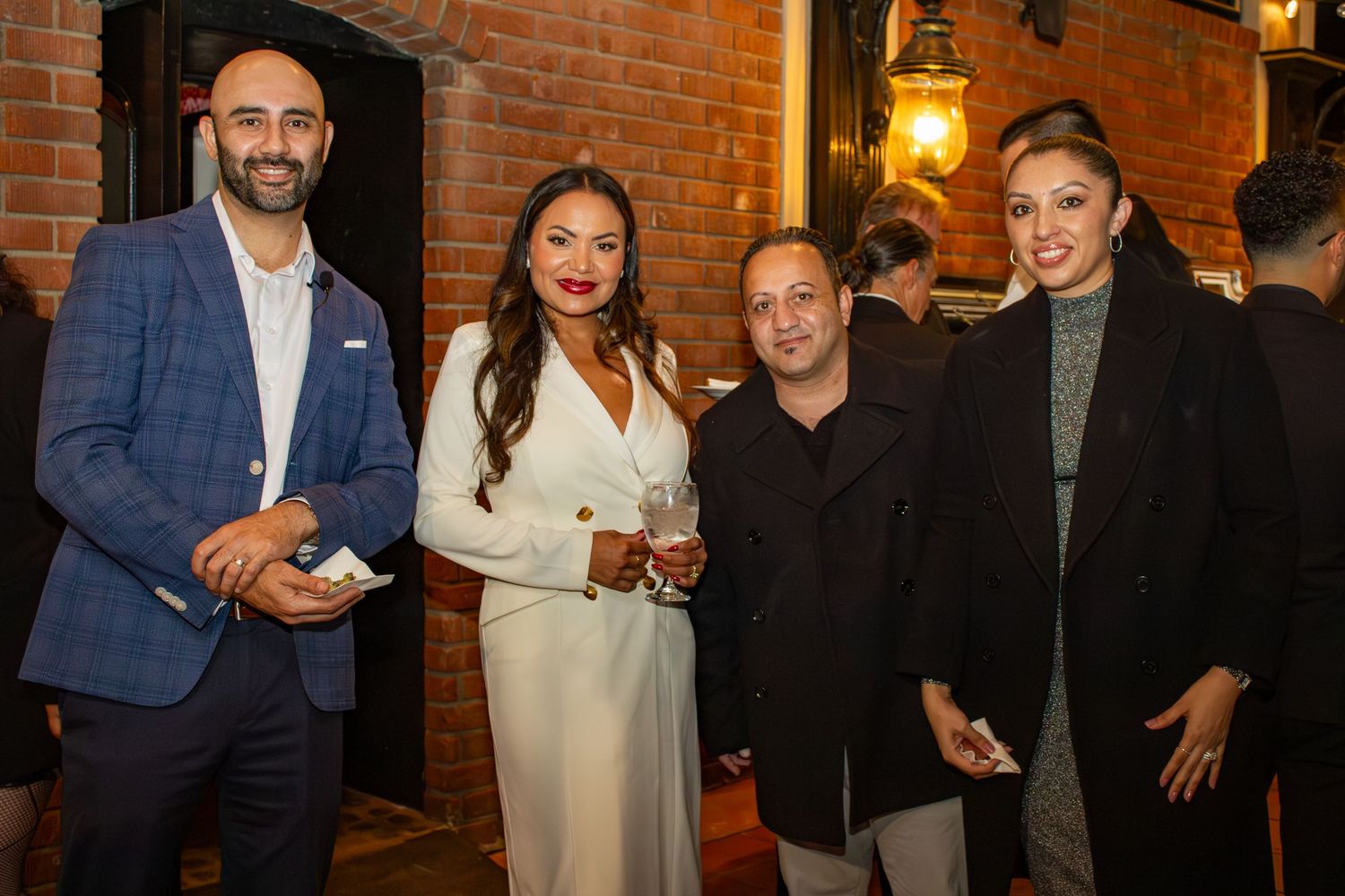 Group of guests in formal attire pose against brick wall at evening reception.