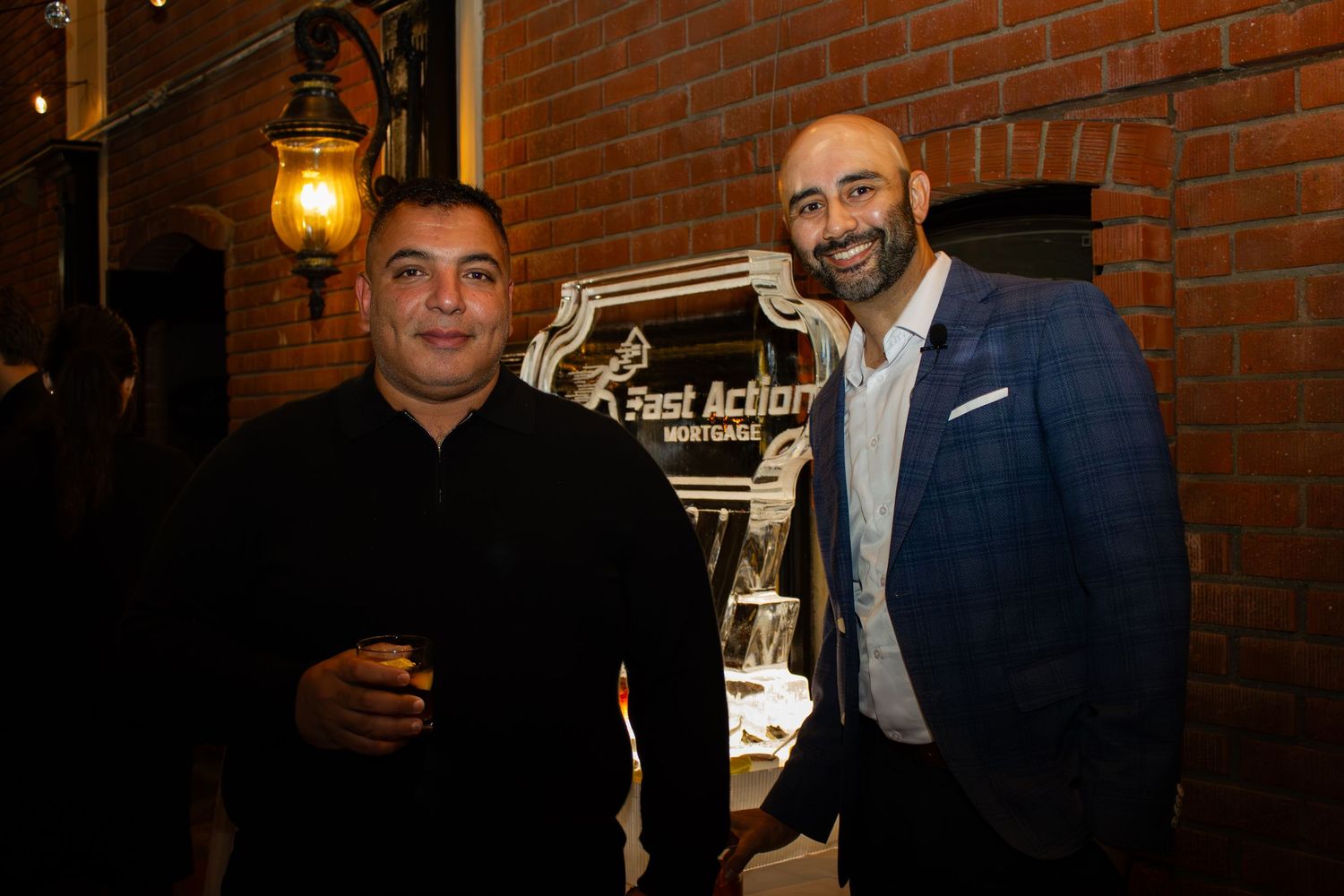 Two guests pose together at an elegant event with a brick wall and vintage lantern in the background.