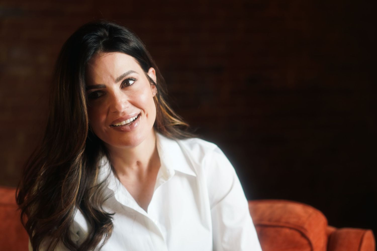 Smiling woman with long dark hair wearing a white button-up shirt, seated on an orange chair in a warmly lit room.