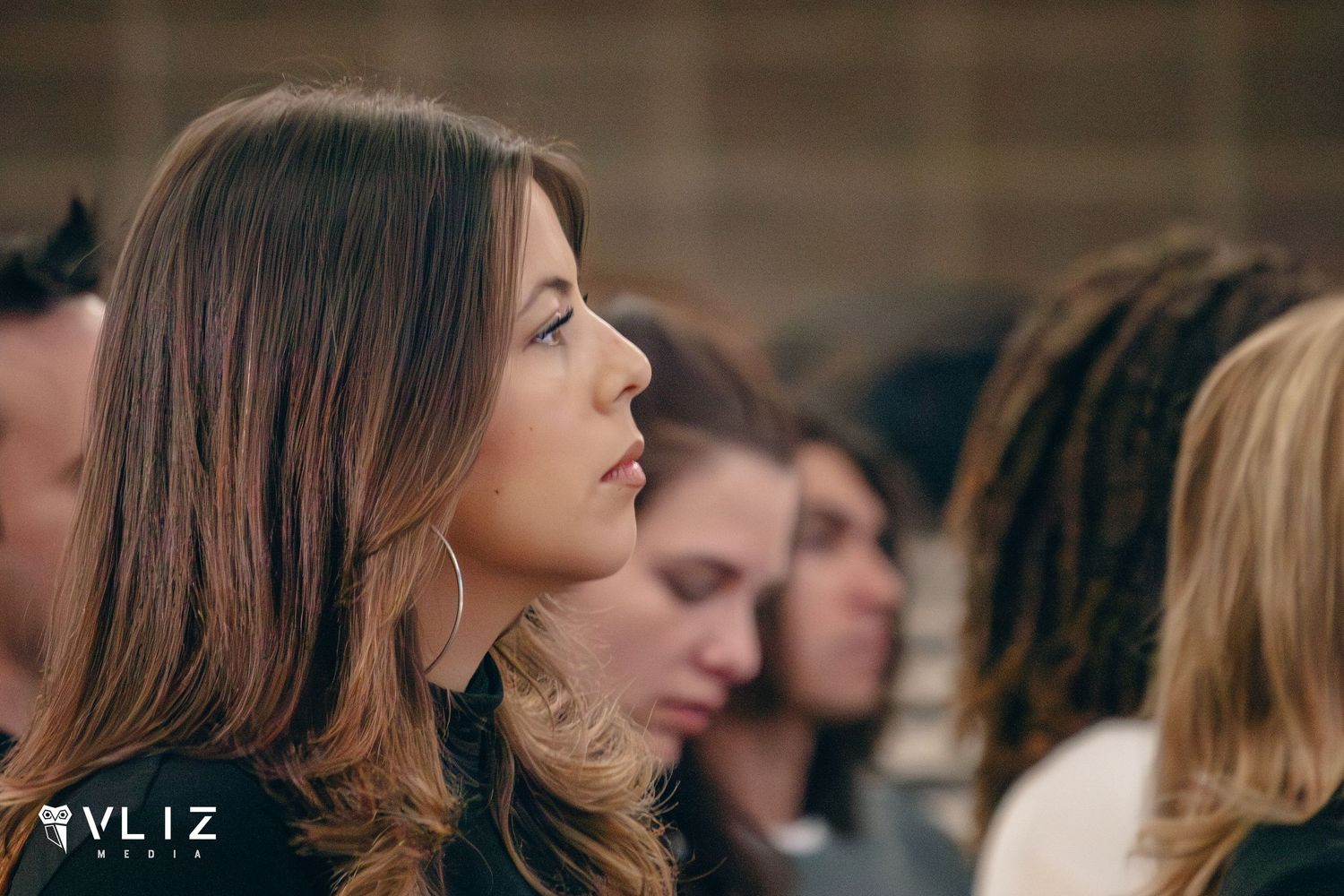 Young woman with long brown hair and hoop earrings gazes upward attentively in a crowd of people.