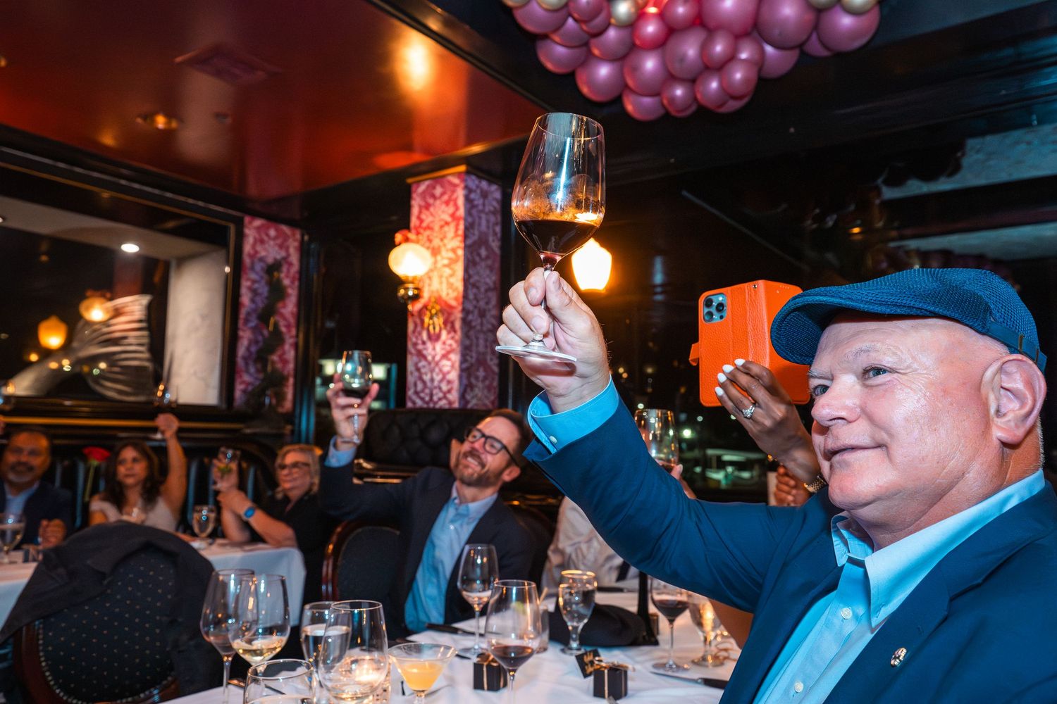 Man in blue cap raises wine glass in toast at elegant restaurant celebration with balloons.