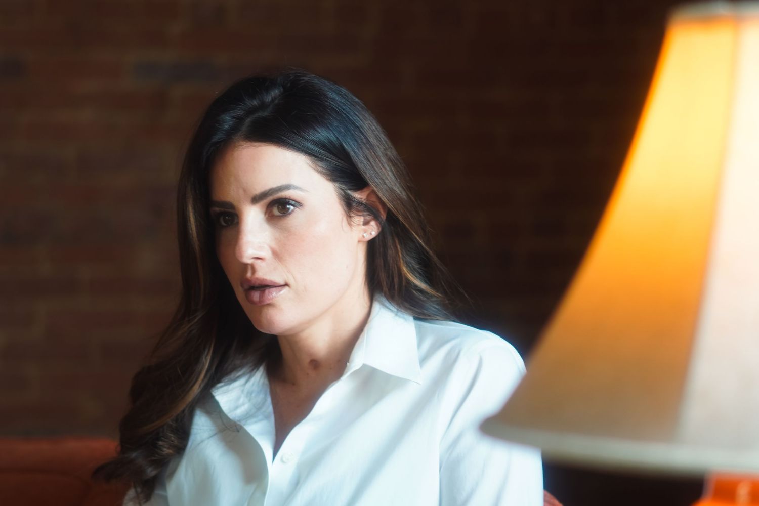 Woman with dark hair in white shirt sits near a glowing lamp against a brick wall background.