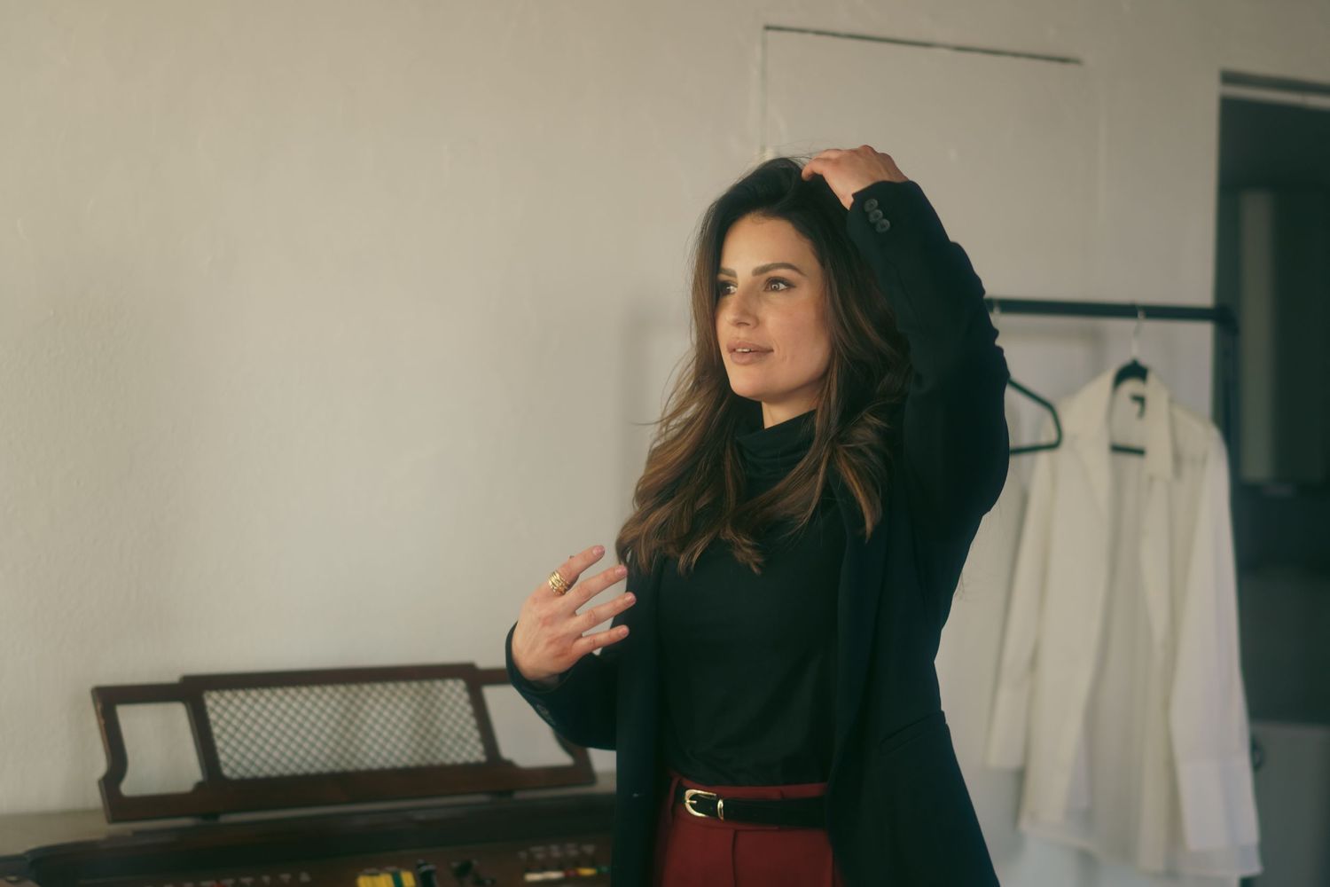 Woman in black blazer and red skirt poses gracefully in a stylish room with clothing rack in background.