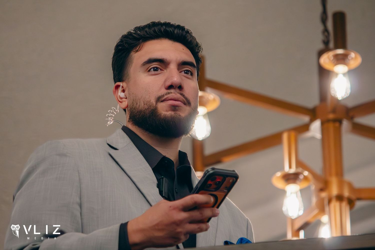 A stylish man in a gray blazer holds a phone, wearing an earring, under warm Edison bulb lighting at an event.