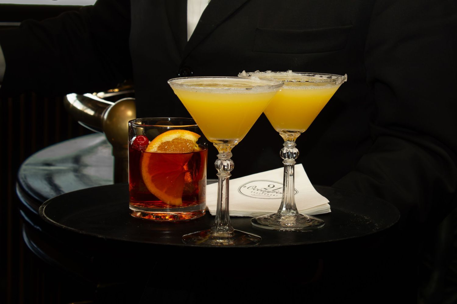 A server holds a tray with two yellow martinis and a classic cocktail garnished with orange and cherry.
