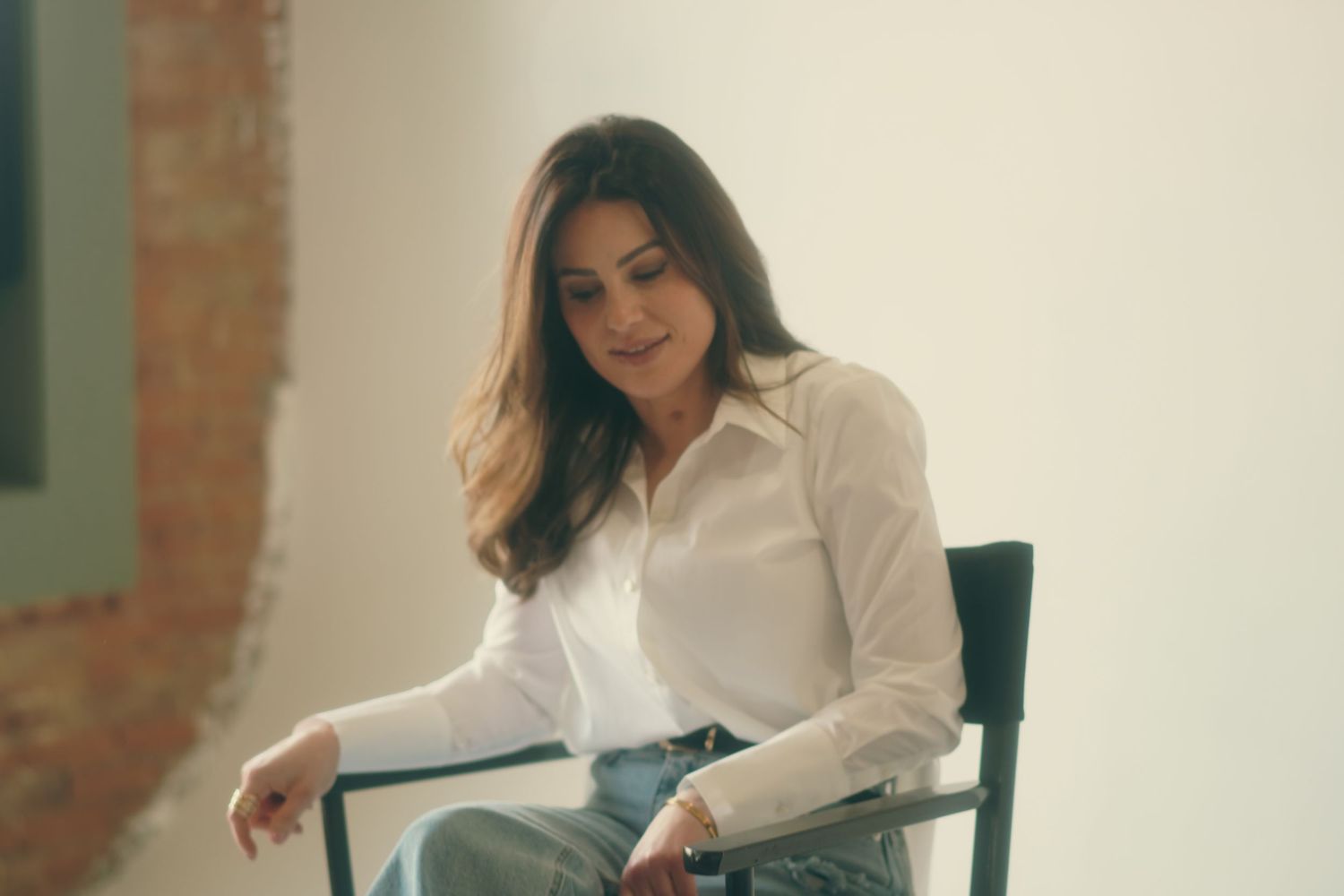 Woman in white shirt and jeans sitting relaxed on a chair, smiling softly in a bright studio setting.
